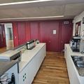 A photograph of the RCR kitchen. There are two white counters facing each other covered in blcck and silver kitchen appliances. The far wall is red and the floor is wooden. There is a silver sink next to the wall.
