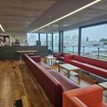 A photograph of the RCR. There are 2 long rows of red sofas facing each other around white coffee tables on the left side of the room. Behind them is a glass window wall. In the background is the RCR kitchen. The floor is wooden.