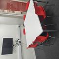 A photograph of meeting room 1. There is a white table in the middle of the room with 6 red chairs around it. A TV screen is on the wall behind the table. The walls are white and there is a glass door.