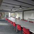 Photograph of the conference room with a long table surrounded by red chairs. There is a large map at the end of the room.