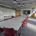 Photograph of the conference room with a long table surrounded by red chairs. There is a large screen at the end of the room.