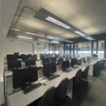 A photograph of the computing lab. There are rows of white desks with 4 computers on each desk in front of a black chair. At the front is a large whiteboard and there are floor to ceiling windows on the far wall.