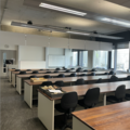 A photograph of the elementary lab. There are 5 rows of wooden desks and black chairs looking at 2 large whiteboards and a lecturing podium with a computer on it. There are large windows on the far wall. A tray of rock sits on one of the desks.