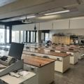 A photograph of the elementary lab. There is a lecturing podium with a laptop looking at 5 rows of wooden desks and black chairs. In the back of the room are large wooden cupboards. There are large windows on the far wall.