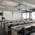 A photograph of the mineralogy lab. There are 4 rows of wooden desks in front of 2 large white boards and a lecturing podium. There are large windows on the far wall. 