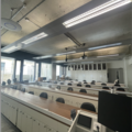  A photograph of the mineralogy lab. The photo is taken facing 5 rows of wooden desks with black chairs behind them. There is the corner of a lecturing podium in the bottom right of the image and you can see a blank laptop screen. There are large windows.