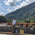 A view from a bridge over a river in a forested valley. A person is standing on the bridge with a bucket and rope.