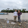 Two people standing on a rocky beach with a bucket and water sample in front of a wide river.