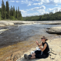 a woman sampling river water, sitting next to a wide river on a plateau of limestone surrounded by forest