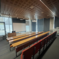 Photograph of Seminar Room 2. There are 4 rows of red chairs with wooden tables in front of them facing a large projector screen at the front of the room. Next to the projector is a podium with a computer on it and floor to ceiling windows. 