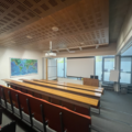 Photograph of seminar room 1. There are four rows of red chairs with wooden desks in front of them facing a large projector screen. A podium is next to the screen and behind is floor to ceiling windows. On the far wall is a world map.