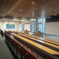 Photograph of the Seminar Rooms. There are four rows of red chairs with brown wooden desks in front of them facing two large projector screens. Behind the screens are floor to ceiling windows. There is a large world map on the far wall.