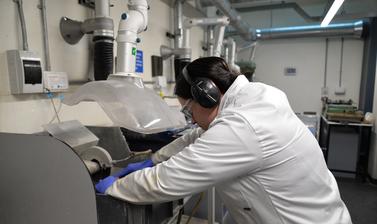 Researcher using a cutting machine to break apart a rock. They are wearing ear defenders and a lab coat and leaning over the machine.