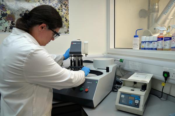 Geofacilities manager Emily polishes a sample using a Struers LaboPol-35 polishing machine while wearing a lab coat and gloves. Bottles of chemicals and other equipment can be seen in the background