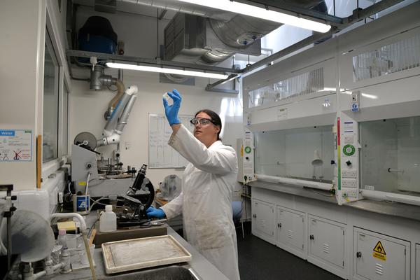 Lab manager Emily holds a thin section up to the light in the thin sections lab. She is wearing a lab coat and gloves. Other equipment including fume cupboards is visible in the background.