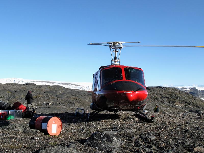 Helicopter at the Isua Supracrustal Belt Photograph of a red helicopter on dark grey rock with the ice sheet in the distance