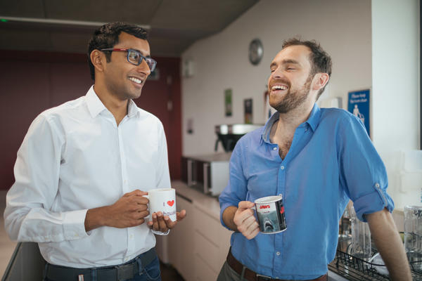 Photograph of two people chatting and laughing over a mug of coffee. They are stood in front of large glass windows.
