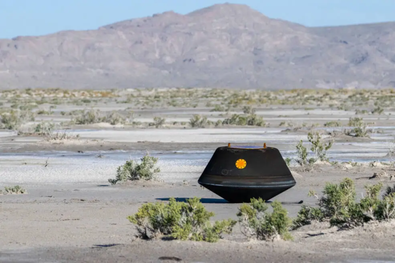 A photograph of the Bennu Sample Return Capsule. It is a large black hexagonal craft and is sat on a large plain with mountains in the background.