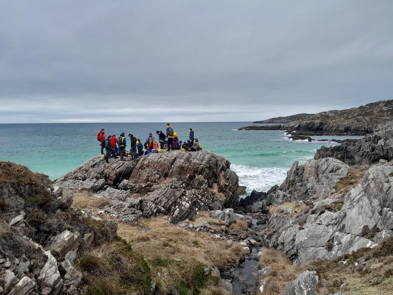 Photograph from a distance of a large group of students stood on a huge rock overlooking a blue-green sea and white beach