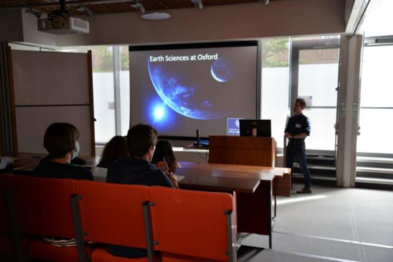 A photograph of a member of staff delivering an introductory presentation at a department open day. They are standing next to a large screen displaying presentation slides