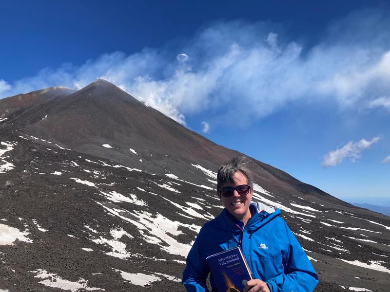 Tamsin Mather Etna Photograph of Tamsin stood on Etna holding a copy of her book, smiling at the camera. She is wearing sunglasses and a blue jacket. The sky is blue and has small white clouds.