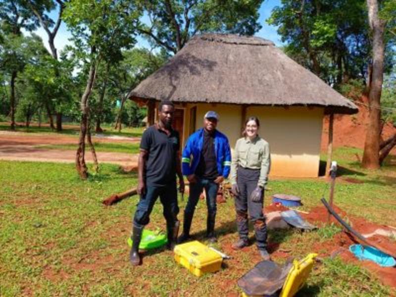Rita Seismics Field Photo Three people standing in front of a little hut with a thatched roof in the middle of some trees in Zambia. On the ground in front of them is some digging equipment. They are dressed in boots and field gear.