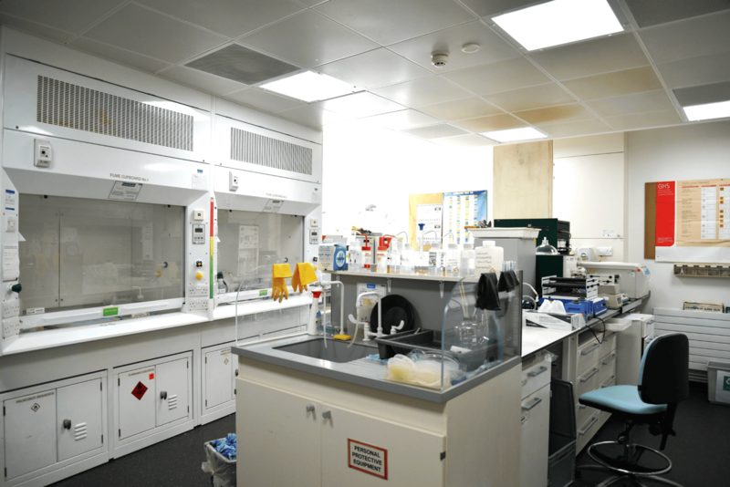 Wet chemistry lab with two fume cupboards on the left, a central sink and workbench, and an empty chair on the right.
