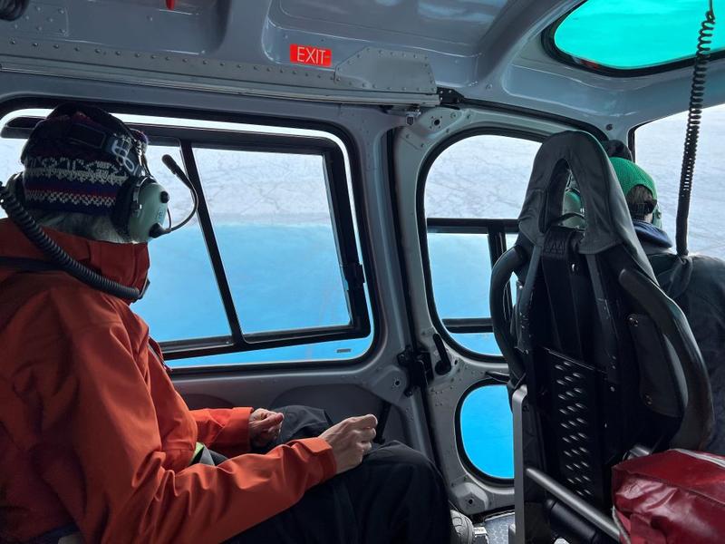 Laura in a Helicopter A photo from inside of a helicopter showing a person in a hat and orange coat wearing a headset and looking out of the window at blue sea to the bottom of the window and ice to the top of the window. Infront of this seat you can see the front seat of the