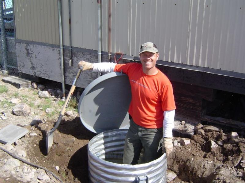 Mike standing ina. corrugated metal tube coming out of the ground. He is holding a large spade. Behind him is a warehouse building and around him is dirt and rubble. 