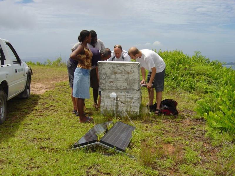 Mike, along with 4 other people, looking into a metal box that is linked to a solar panel. They are in a grassy field with a white car parked next to them.