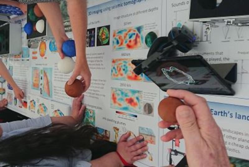 A photo of a table covered in posters about seismometry and three hands over the table holding 3D globe models