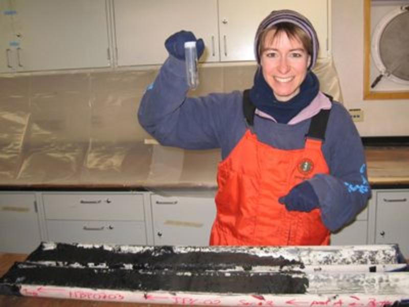 A photo of Ros wearing snow gear holding up a test tube standing behind a table with her experiment in it. She is inside a wooden hut.