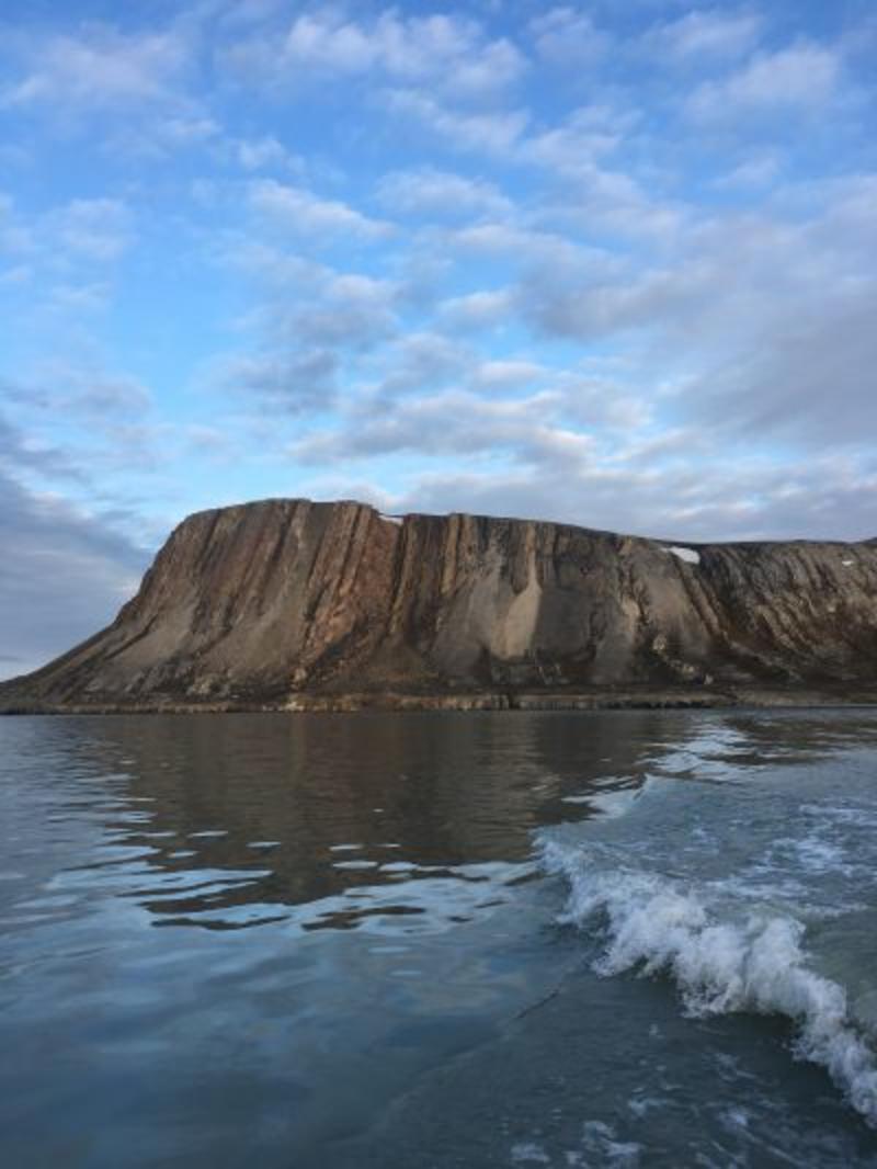Svanbergfjellet Formation A tall brown cliff rising from the sea. It has vertical lines going down it. The sky in blue and dotted with clouds.