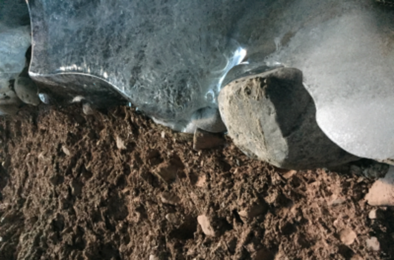 Ice in the top of the image meets brown rocky ground at the bottom of the image. Pebbles of various sizes are located at the interface.