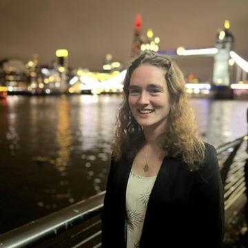 Carys standing incfront of a river. She is wearing a white top and a black blazer, her hair is down in front of her shoulders. You can see a bridge and lights of the city in the background.