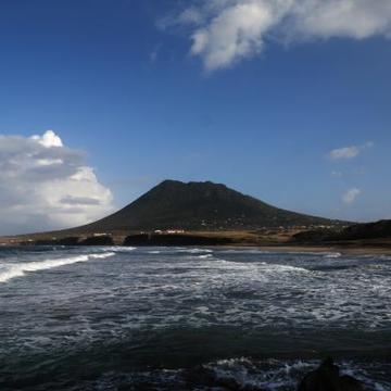 The Quill Volcano Image shows a volcano. The photo is taken across a sandy bay looking over the sea at the volcano.
