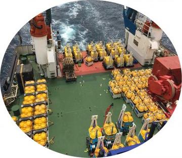 Circular image looking down at the back of a ship. The deck is green and is covered in yellow containers. There is a person at the end of the ship standing on a lader next to a red and white tower.
