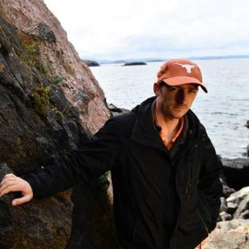 Richard Palin standing next to a big rock with the sea in the background. He is wearing a brown coat and a red baseball cap.