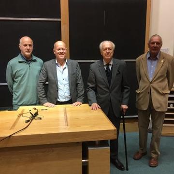 Four men wearing suits standing behind a wooden desk in front of a black chalk board