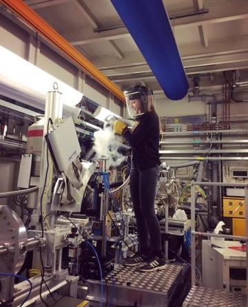 Rosalie in the Lab A scientist wearing a face protection shield stands on a metal stool pouring some steaming gas into a machine. They are in a lab filled with metal equipment and machines.