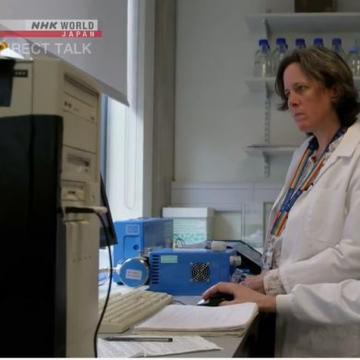 Tamsin wearing a white lab coat and blue gloves standing in a lab looking at a computer screen.