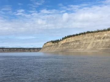 Mackenzie River Sed rocks A wide blue river int he foreground, on the far bank are some yellow-beige cliffs with fir trees all along the top. The sky is blue and filled with fluffy clouds.