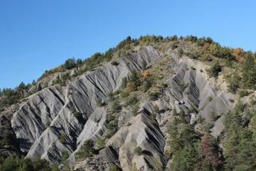 Erosion in South of France Photo looking up a grey mountain soparsely covered in green fir trees. The rocks are jagged and form ridges going up the mountain. Above it is a clear blue sky.