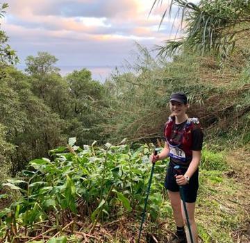 Claire on a hike. She is wearing a hiking backpack and carrying walking poles. She is standing amongst lots of vegetation on a mountain with the sea in the far distance behind her