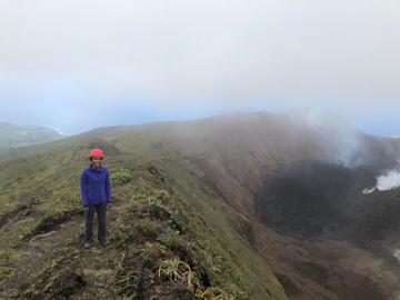 Michal standing on the edge of a volcano crater. They wears a blue coat and orange hard hat. The ground below her is grassy and you can see steam rising from the crater behind her 
