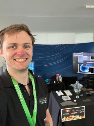 A selfie of James wearing a black top and a green landyard in front of a display table.