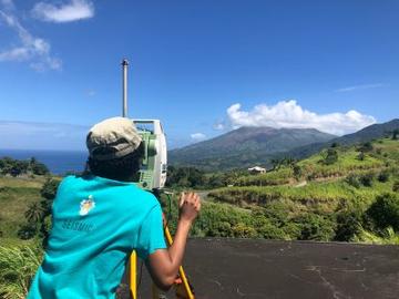 A photo taken from behind Michal as she looks into a monitoring device at a vegetation covered volcano in the distance 