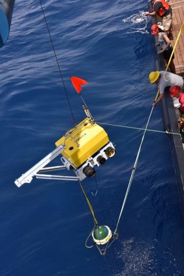 Image shows a yellow container being lowered off the side of a large ship into the open ocean. There are people wearing yellow and red hard hats on the side of the ship.
