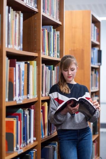 Photograph of a students reading in the library. They are stood leaning against a large wooden bookcase of books.