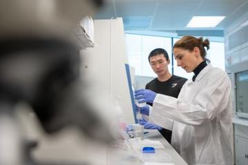 Image of Julie and a male colleague in the lab. She is wearing a white lab coat and blue gloves and is conducting an experiment. the foreground is a blurry unfocused microcope.